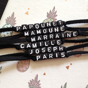 Op de afbeelding: Zes zwarte kralenarmbanden met witte letters die de namen "Papounet", "Mamoune", "Marraine", "Camille", "Joseph" en "Paris" spellen.
