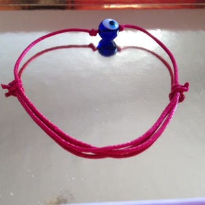 Puede incluir: Un brazalete de cuerda rosa con un dije azul de ojo turco.