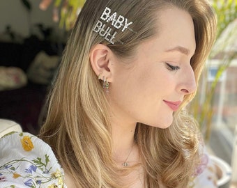 May include: A woman with long blonde hair wearing a white floral blouse with a pair of silver hair clips that say "BABY BULL" in rhinestones.