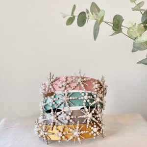May include: Four headbands with a starburst design. The headbands are in pink, teal, gray, and yellow. Each headband is decorated with silver rhinestones and white pearls.
