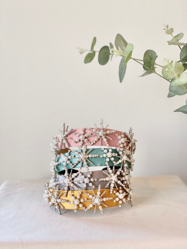 May include: Four headbands with a starburst design. The headbands are in pink, teal, gray, and yellow. Each headband is decorated with silver rhinestones and white pearls.