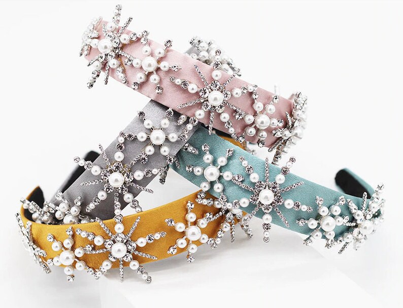 May include: Three velvet headbands with snowflake embellishments. The headbands are in pink, gray, and yellow. Each headband is decorated with silver rhinestones and white pearls.