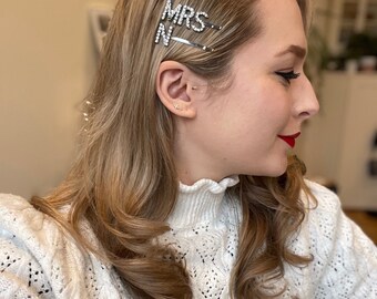 May include: A woman with long blonde hair wearing a white sweater with a ruffled collar. She has two hair clips in her hair that say "MRS" and "N" in rhinestones.