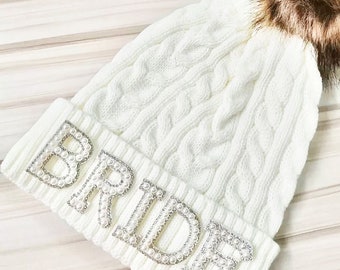 May include: White knitted beanie with a faux fur pom pom and the word "BRIDE" spelled out in pearl-like embellishments.