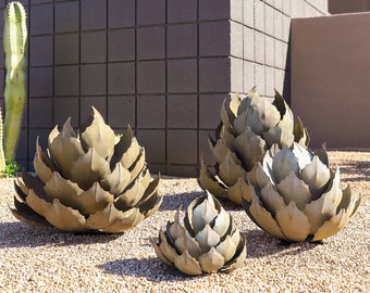 May include: Four metal sculptures of agave plants in various sizes, arranged on a bed of small brown pebbles.