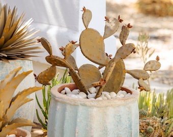 May include: A metal cactus sculpture in a light blue ceramic pot with white stones. The cactus is rusty brown and has a realistic shape.