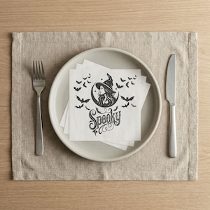 May include: A stack of white paper napkins featuring a black graphic print of a witch wearing a hat, a crescent moon, and several bats, with the word Spooky written in a decorative script font, centered on a light gray plate with a fork and knife.