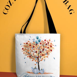 May include: A white tote bag with black handles featuring a graphic of a tree with orange, red, and blue leaves, two blue pumpkins at the base, and the text "Autumn Is My Happy Place" at the bottom. The words "COZY FALL TOTE BAG" appear above.