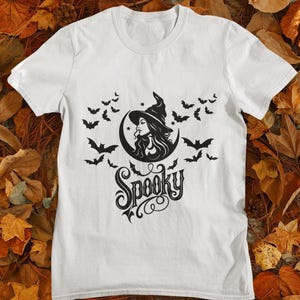 Może przedstawiać: Biały t-shirt z krótkim rękawem z czarnym nadrukiem graficznym przedstawiającym czarownicę w spiczastym kapeluszu na tle półksiężyca, otoczoną przez latające nietoperze. Słowo Spooky jest nadrukowane czcionką pod główną grafiką.
