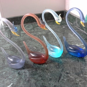May include: Five glass swan figurines in different colors, including blue, light blue, pink, and clear. The swans are arranged in a row on a green marble surface.