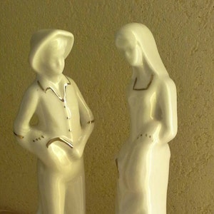 May include: A pair of white porcelain figurines depicting a man and a woman. The man is wearing a hat and a jacket, while the woman is wearing a long dress. Both figurines have gold accents.