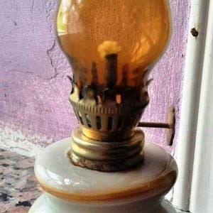 May include: A small, amber glass oil lamp with a brass burner and a white marble base. The lamp is sitting on a granite countertop.