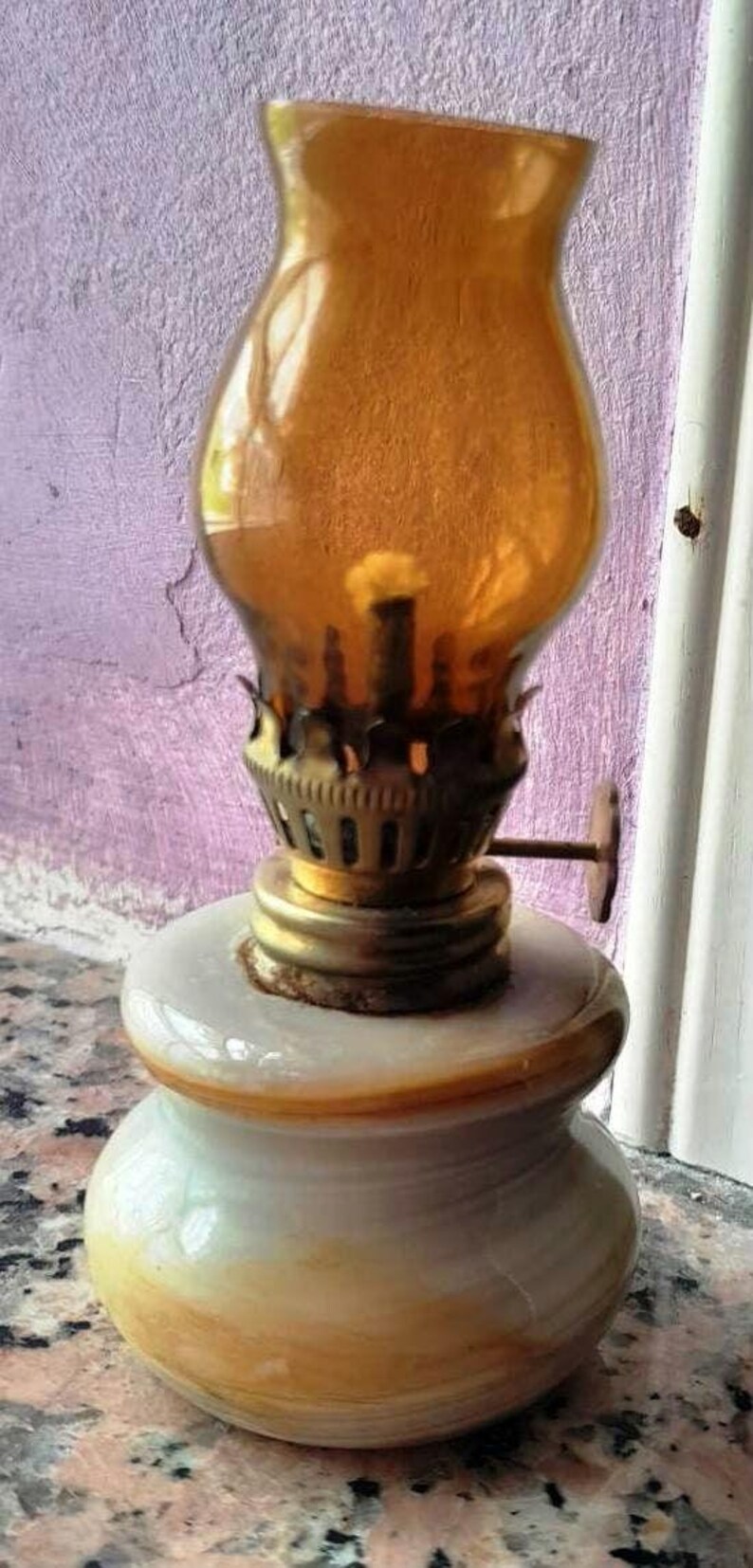 May include: A small, amber glass oil lamp with a brass burner and a white marble base. The lamp is sitting on a granite countertop.