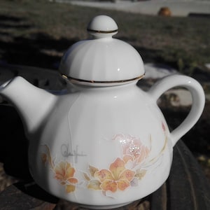 Op de afbeelding: Een witte theepot van keramiek met een bloemenmotief. De theepot heeft een gouden rand rond de deksel en de tuit. De bloemen zijn roze, geel en oranje.