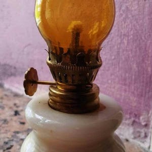May include: A small, white, ceramic oil lamp with a gold-colored metal wick holder and a clear amber glass chimney.