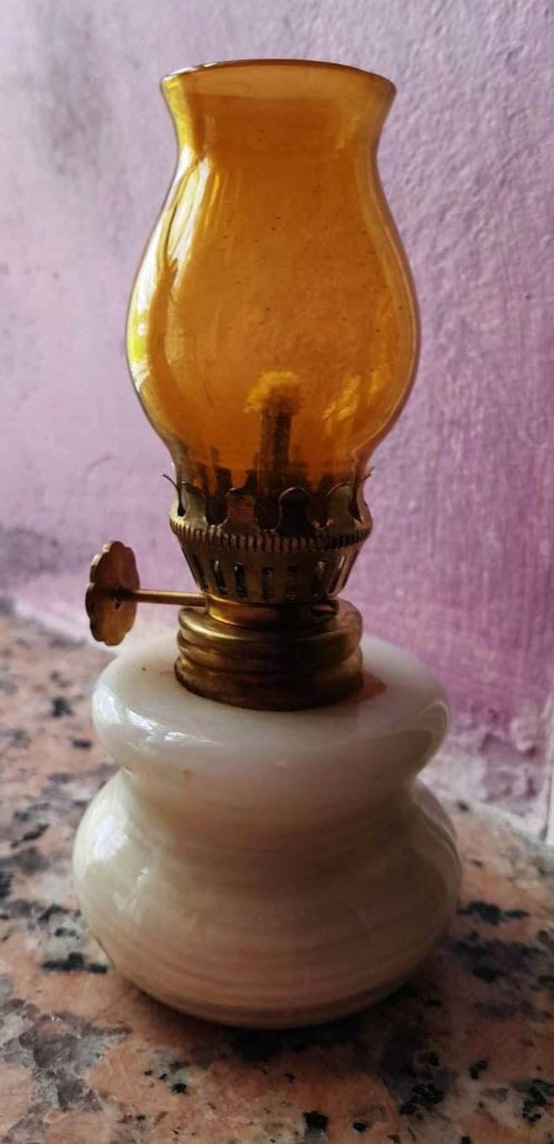May include: A small, white, ceramic oil lamp with a gold-colored metal wick holder and a clear amber glass chimney.