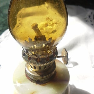 May include: A small, antique oil lamp with a white marble base and a clear glass chimney. The lamp has a brass wick holder and a small metal knob on the side.