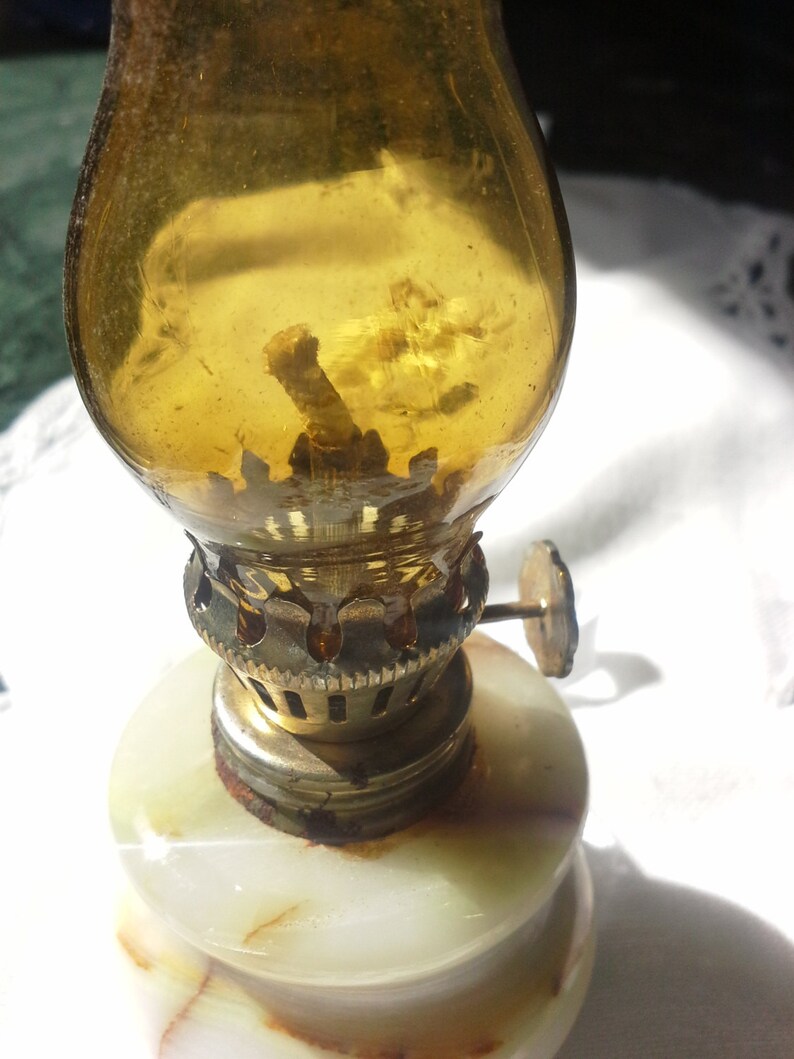 May include: A small, antique oil lamp with a white marble base and a clear glass chimney. The lamp has a brass wick holder and a small metal knob on the side.