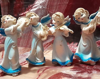 Puede incluir: Cuatro figuritas de ángeles de porcelana blanca tocando instrumentos musicales. Los ángeles están vestidos con túnicas blancas con ribetes azules y tienen el pelo rubio. Los instrumentos incluyen un pandero, una lira, un violín y un laúd.