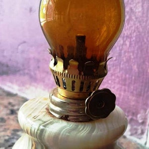 May include: A small oil lamp with a white marble base and a clear amber glass chimney. The lamp has a brass wick holder and a brass knob on the side.