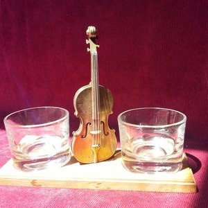 May include: Two clear glass shot glasses sit on a wooden base with a gold-colored miniature cello in the center.