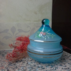 May include: A blue glass jar with a lid, decorated with gold trim and painted flowers. The jar has a frosted finish and is perfect for storing small items.