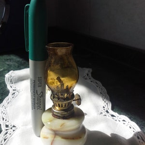May include: A small, decorative oil lamp with a marble base and a glass chimney. The lamp is made of white marble with brown veins and has a gold-colored metal top. The glass chimney is amber-colored and has a small metal handle.