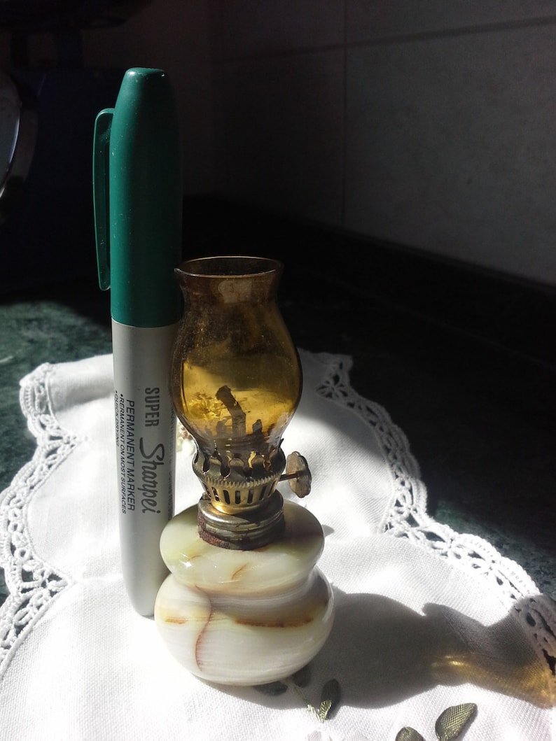 May include: A small, decorative oil lamp with a marble base and a glass chimney. The lamp is made of white marble with brown veins and has a gold-colored metal top. The glass chimney is amber-colored and has a small metal handle.