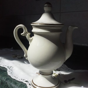 May include: Cream-colored ceramic teapot with a lid and a curved handle. The teapot has gold trim around the base, lid, and spout. It sits on a pedestal base and is placed on a white lace doily.