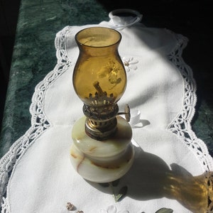 Puede incluir: Una pequeña lámpara de aceite de vidrio ámbar con una base de mármol blanco. La lámpara está sentada sobre un encaje blanco de ganchillo.