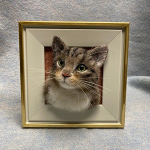May include: A framed needle felted portrait of a gray tabby cat with green eyes. The cat is looking to the right of the frame. The frame is gold with a white mat.