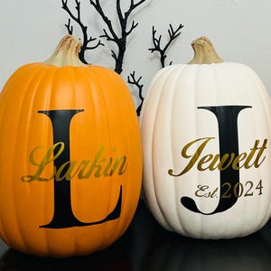 May include: Two decorative pumpkins, one orange and one white, with black letter and name designs. The orange pumpkin has a large black "L" with "Larkin" in gold script. The white pumpkin has a large black "J" with "Jewett Est. 2024" in gold script.