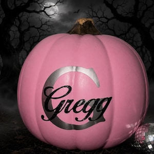 May include: A round, smooth, light pink pumpkin featuring a large, silver-colored letter C as a background for the black, glittery, cursive text Gregg. The pumpkin has a textured, dark brown stem at the top and a small, reflective disco ball.