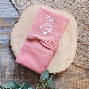 Puede incluir: Una manta de muselina rosa con un corazón blanco y el texto "Notre Jolie Famille" en ella. La manta está atada con una cinta rosa.