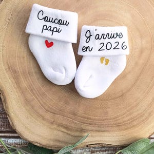 Op de afbeelding: Een paar witte babysokken met geborduurde berichten. E&eacute;n sok zegt "Coucou papi" met een rood hart. De andere sok zegt "J'arrive en 2026" met gouden voetafdrukken.
