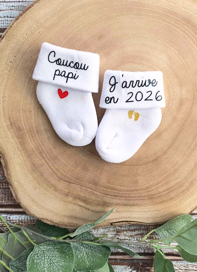 Op de afbeelding: Een paar witte babysokken met geborduurde berichten. E&eacute;n sok zegt "Coucou papi" met een rood hart. De andere sok zegt "J'arrive en 2026" met gouden voetafdrukken.