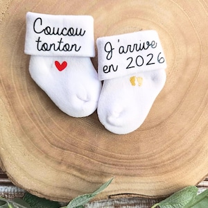 Op de afbeelding: Twee witte babysokken met geborduurde tekst. E&eacute;n sok zegt "Coucou tonton" met een rood hart. De andere sok zegt "J'arrive en 2022" met gouden voetafdrukken.