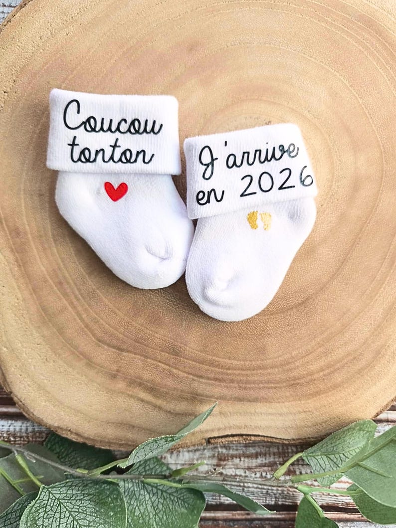 Op de afbeelding: Twee witte babysokken met geborduurde tekst. E&eacute;n sok zegt "Coucou tonton" met een rood hart. De andere sok zegt "J'arrive en 2022" met gouden voetafdrukken.