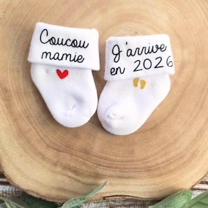 Op de afbeelding: Twee witte babysokken met geborduurde tekst. E&eacute;n sok zegt "Coucou mamie" met een rood hart. De andere sok zegt "J'arrive en 2026" met gouden voetafdrukken.