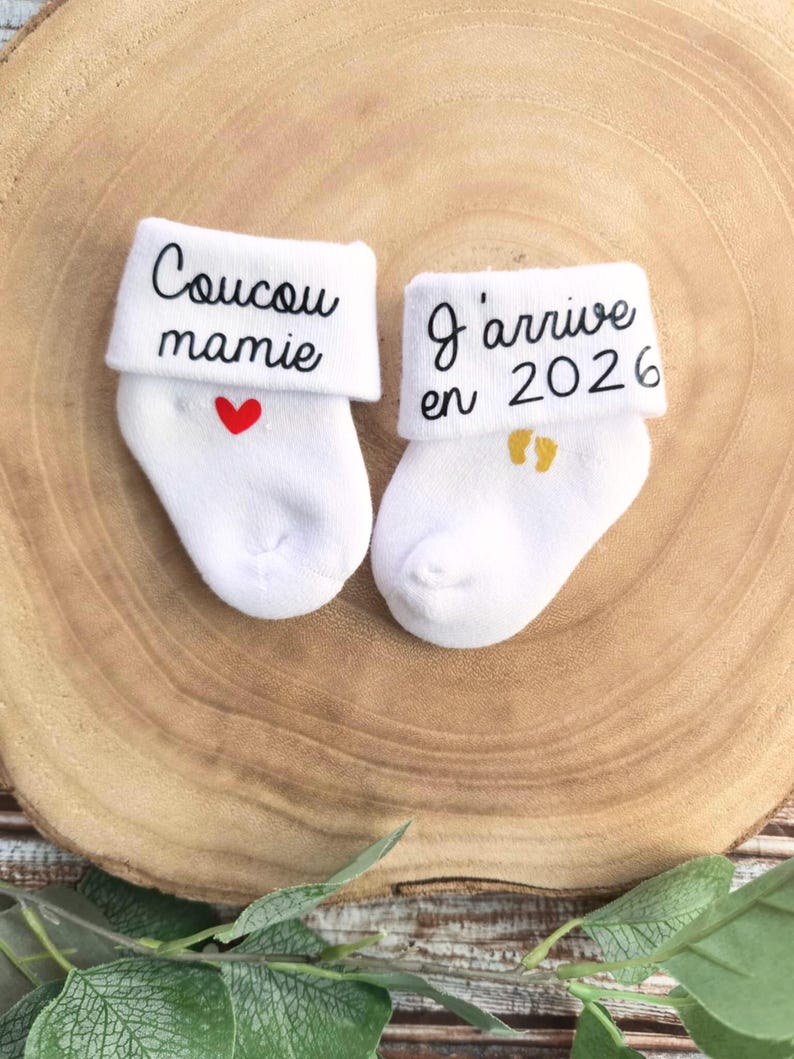 Op de afbeelding: Twee witte babysokken met geborduurde tekst. E&eacute;n sok zegt "Coucou mamie" met een rood hart. De andere sok zegt "J'arrive en 2026" met gouden voetafdrukken.