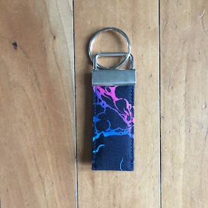 May include: A black fabric keychain with a silver ring and a colorful abstract design. The design features pink, blue, and purple swirls on a black background.
