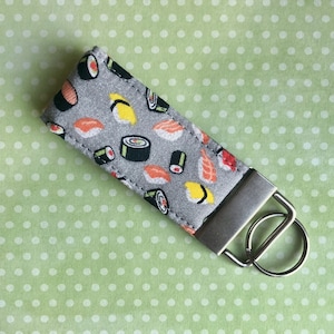 May include: A gray fabric keychain with a silver key ring. The fabric features a repeating pattern of colorful sushi rolls.