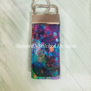 May include: A colorful key chain with a silver key ring and a fabric strap with a blue, green, pink, and yellow abstract pattern. The fabric strap has the text "StampedNStitched.Etsy.com" printed on it.