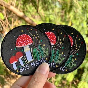 Peut inclure: Trois patchs thermocollants brodés noirs avec un motif de champignon rouge et blanc et le texte "See Magic" en blanc. Les patchs sont sur un fond vert.