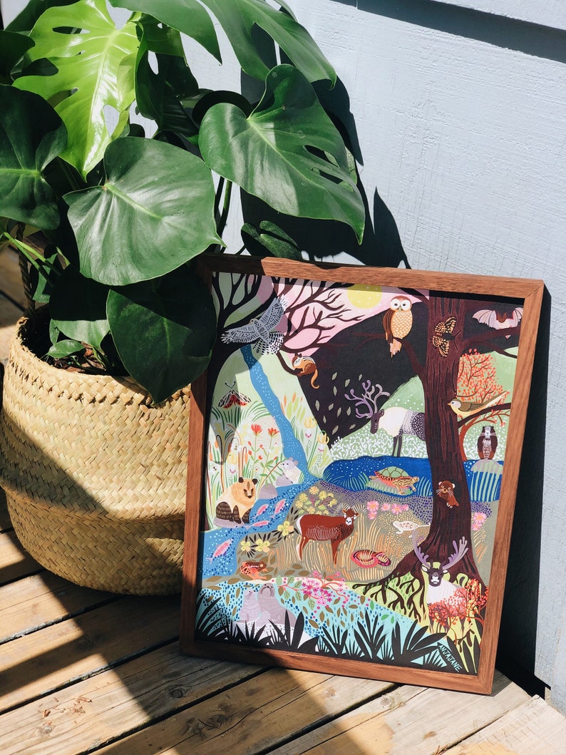 K&ouml;nnte beinhalten: Ein rechteckiger Holzrahmen mit einer farbenfrohen Illustration einer Waldszene mit verschiedenen Tieren wie einer Eule, einem Hirsch, einem B&auml;ren und einer Schildkr&ouml;te, umgeben von B&auml;umen, Blumen und einem Bach, mit der Signatur AMJANE in der Ecke.
