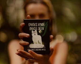 May include: A rectangular oracle deck box with a black background featuring a white wolf illustration, green palm leaf accents, and the text Earth's Hymn Oracle Deck by Alia Jane & Melanie Aryze printed in white lettering on the front cover.