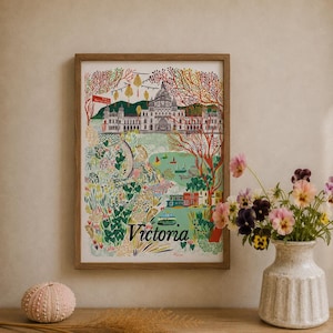 May include: A framed art print featuring a colorful illustration of Victoria, British Columbia, with the Parliament Buildings, Butchart Gardens, and a ferry. The word Victoria is printed in black at the bottom of the white-bordered print.