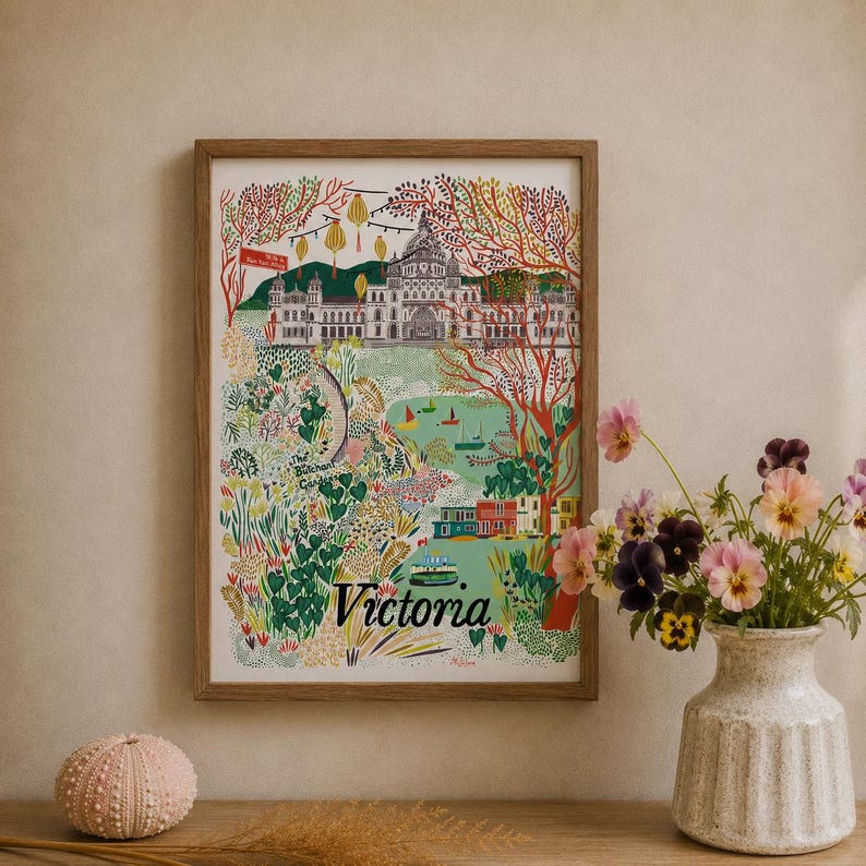 May include: A framed art print featuring a colorful illustration of Victoria, British Columbia, with the Parliament Buildings, Butchart Gardens, and a ferry. The word Victoria is printed in black at the bottom of the white-bordered print.