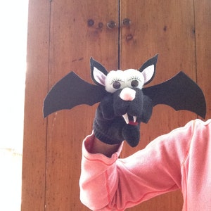 May include: A black and white bat puppet with a pink nose and large eyes. The puppet is being held by a person wearing a pink shirt.
