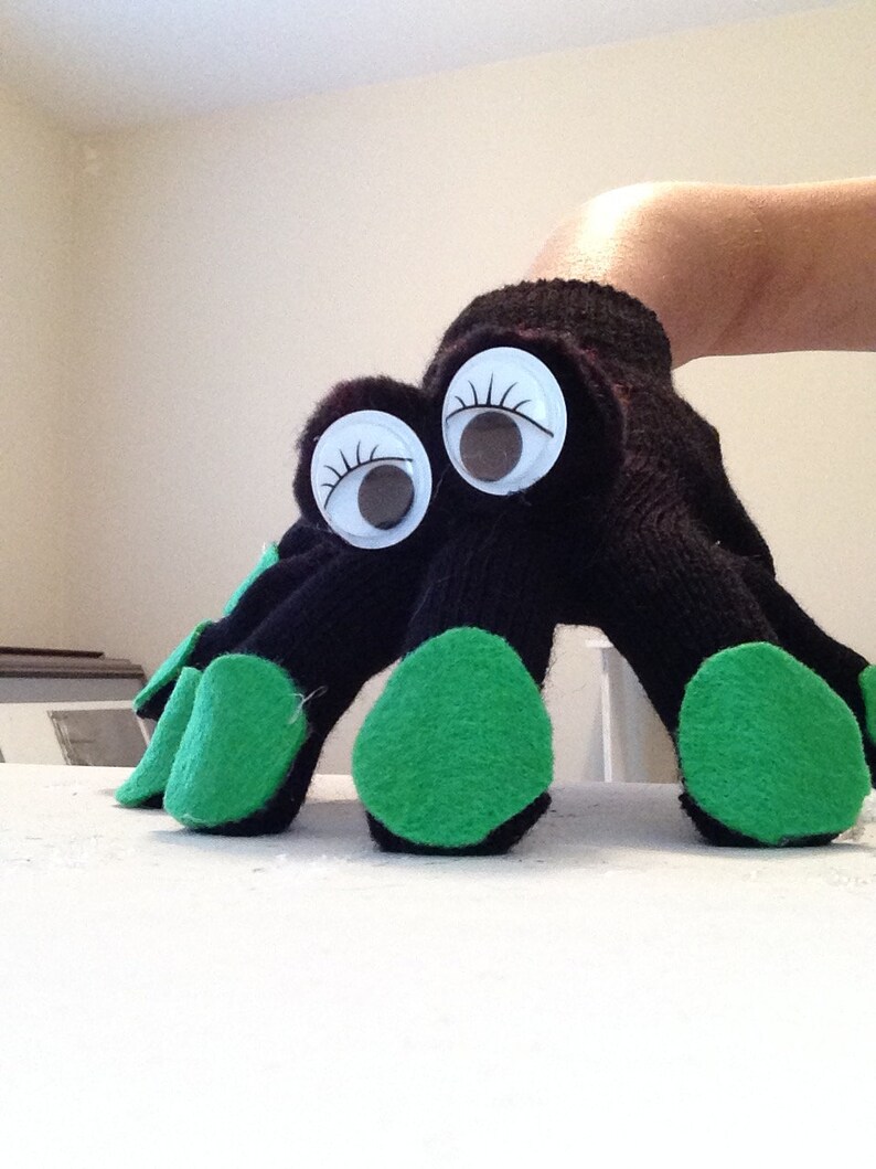 May include: A black sock puppet with large googly eyes and green felt accents on the fingers. The puppet is designed to resemble a creature or monster, with the fingers acting as legs or claws.